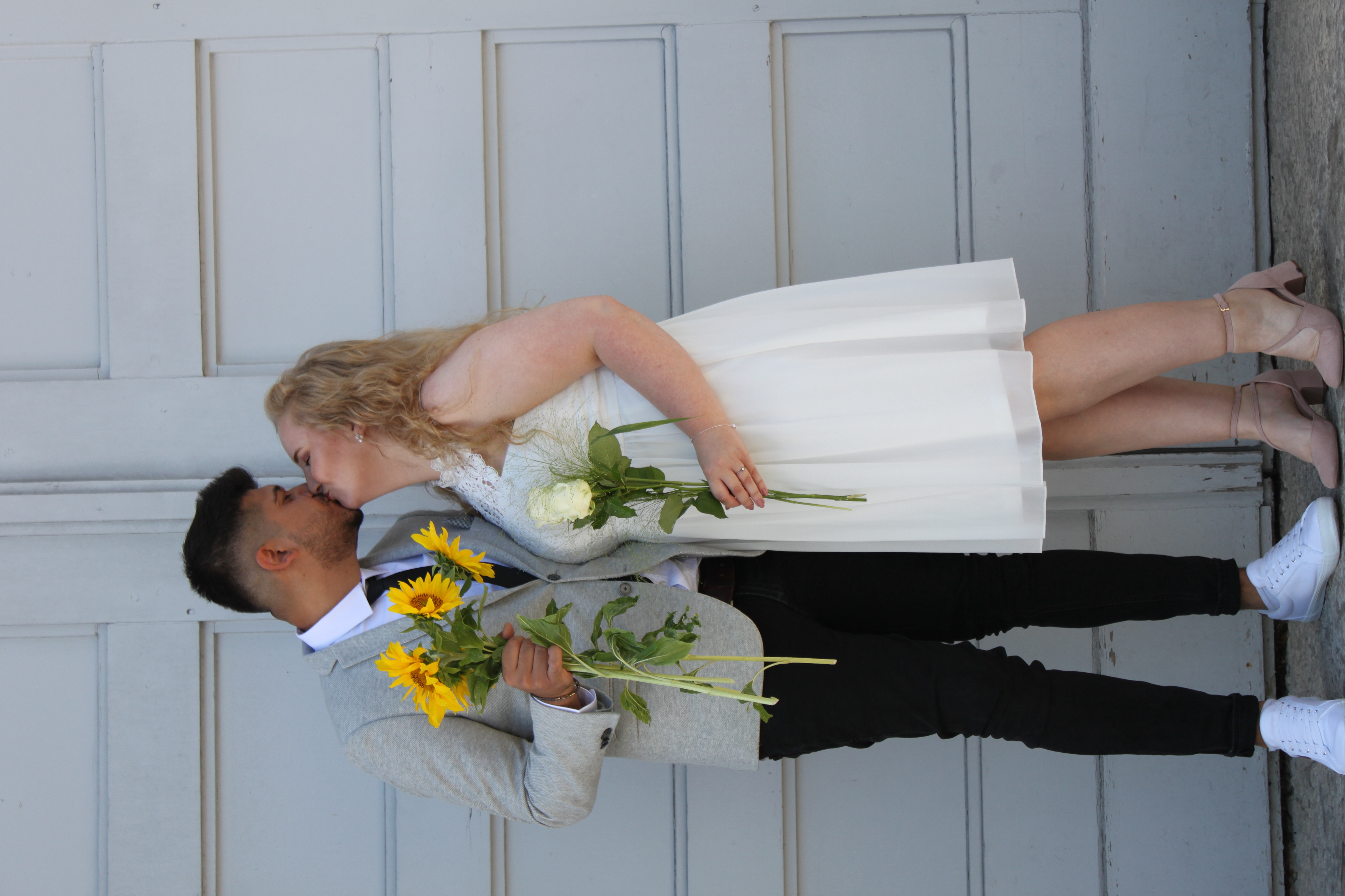 Manuela und Samer an ihrem Hochzeitstag – ein verliebtes Brautpaar, das sich mit Sonnenblumen und weißen Rosen küsst. Sie haben Mitten ins Herz als emotionales Hochzeitsgeschenk erhalten – ein einzigartiges, personalisiertes Geschenk mit Sprachnachrichten und individuellen Podcastfolgen von Familie und Freunden. Das perfekte Hochzeitsgeschenk für unvergessliche Erinnerungen, ideal für Trauzeugen, Geschwister und beste Freunde, die dem Brautpaar eine besondere Freude machen möchten.