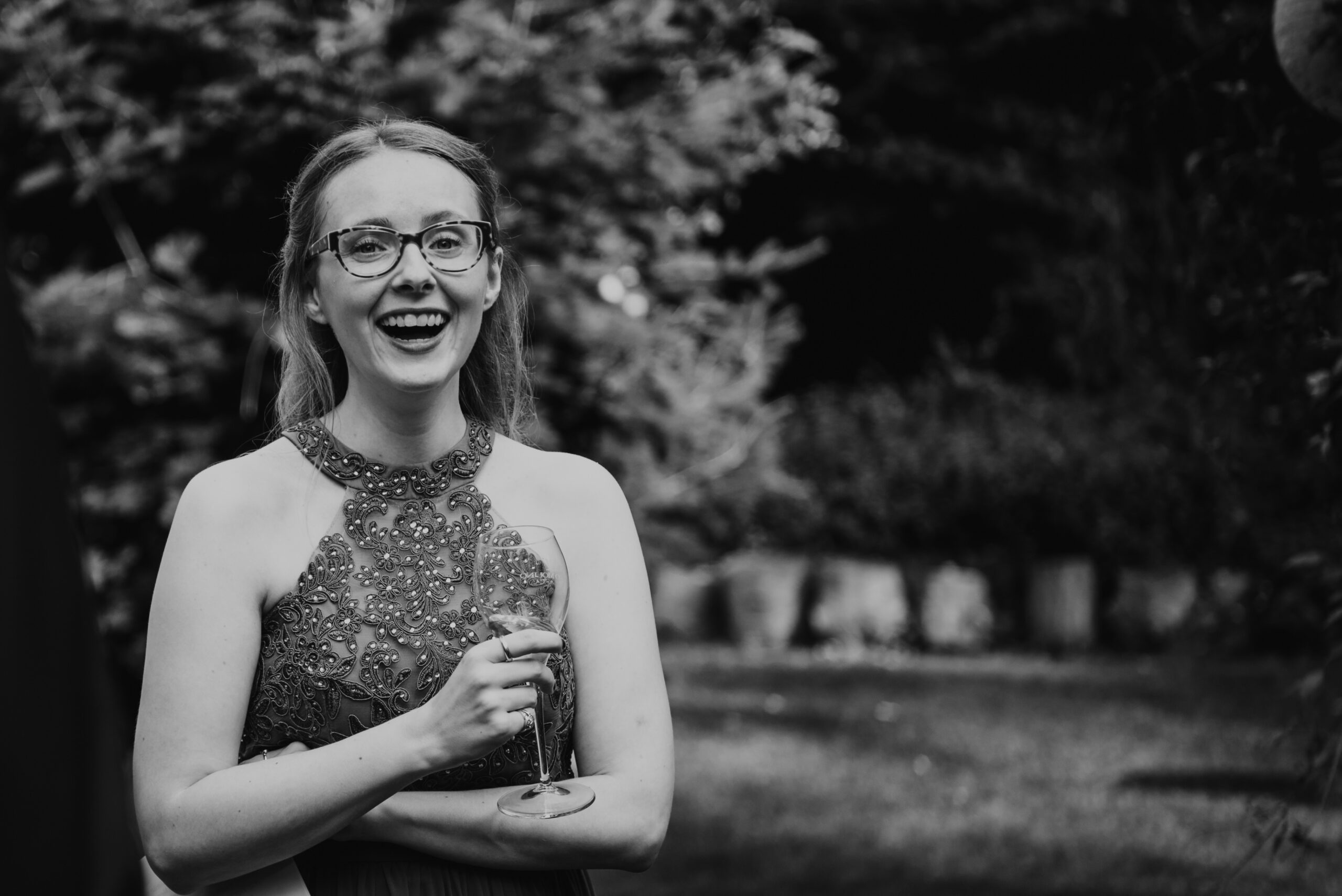 Liza Kummrow, Gründerin von Mitten ins Herz - im eleganten, bestickten Kleid. Sie trägt eine Brille, hält ein Sektglas in der Hand und strahlt Freude aus. Im Hintergrund ein Garten mit Bäumen und Büschen.